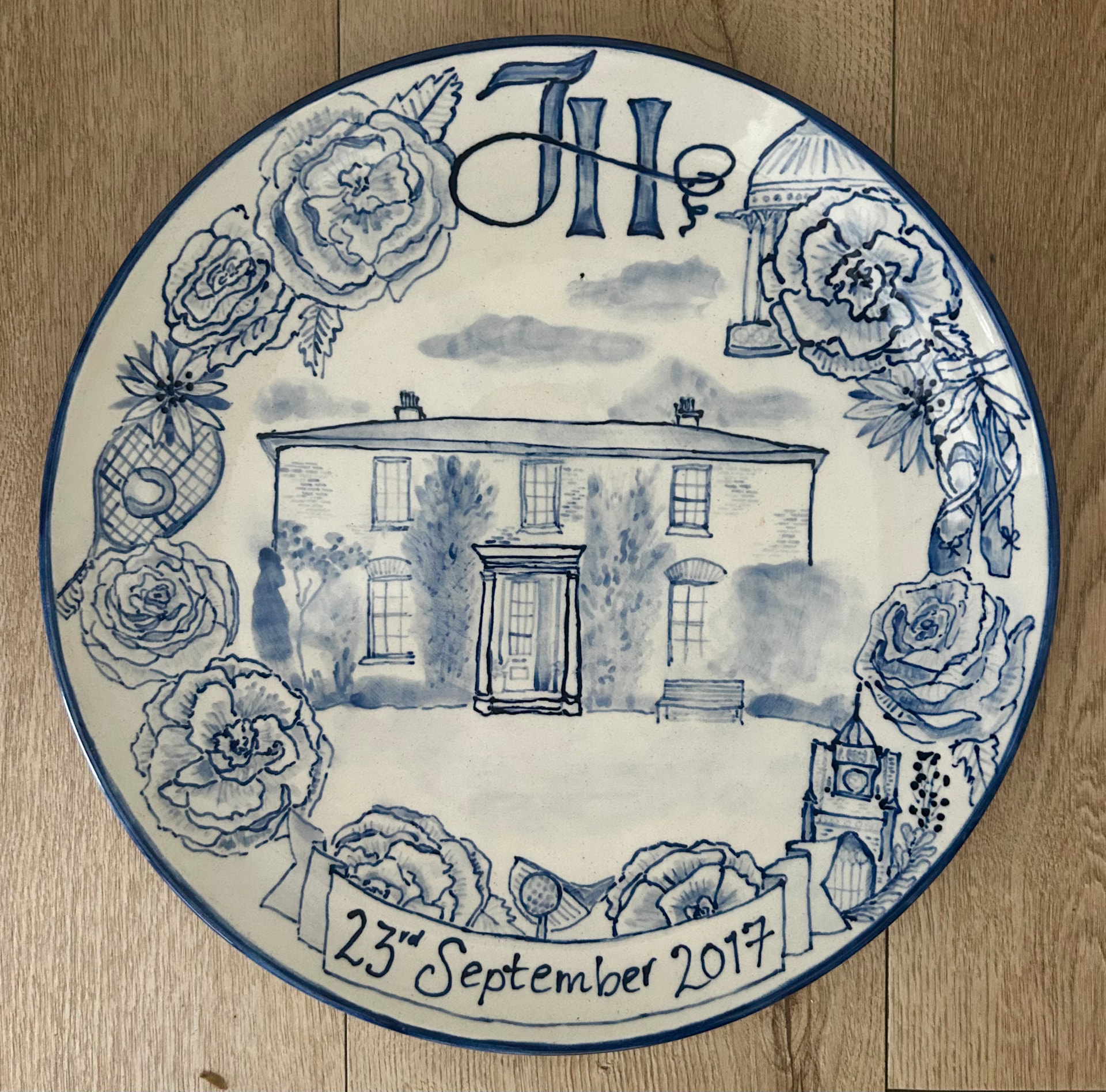 Hand-painted blue and white celebration plate for Hannah and James with central family home, tennis racket, ballet shoes and roses